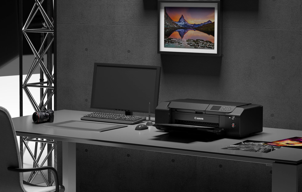 A desk in a studio with a monitor, Canon printer, camera and some prints on it